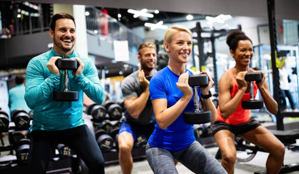 Entraînement en Groupe : Fitness et Musculation avec Haltères Quatre personnes souriantes s'entraînent avec des haltères dans une salle de sport, effectuant des squats.