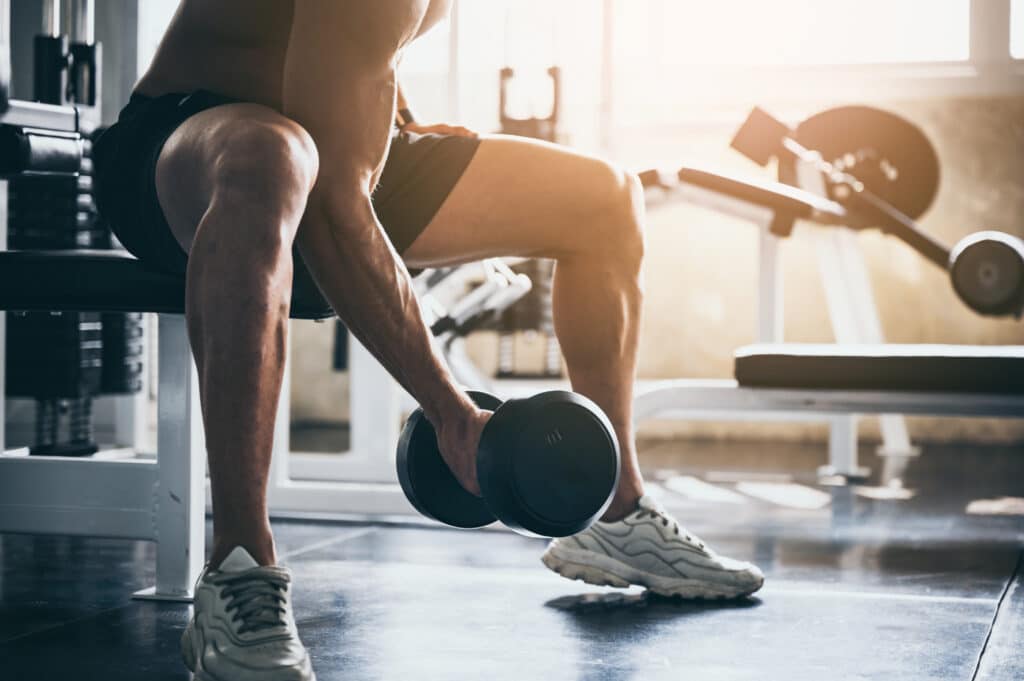 Musculation : Homme puissant soulève des haltères à la gym Homme musclé sans chemise assis sur un banc de gym, soulevant un haltère. Lumière chaude et équipement de fitness en arrière-plan.